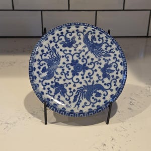 May include: Round, blue and white ceramic plate with a phoenix and floral design. The plate is displayed on a black metal stand. The plate's diameter is approximately 6 inches.
