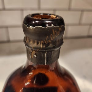Pode incluir: Close-up de uma garrafa de vidro castanho com uma tampa castanha escura. A tampa tem uma superf&iacute;cie texturizada e uma inscri&ccedil;&atilde;o dourada. A garrafa est&aacute; parcialmente cheia de um l&iacute;quido escuro.