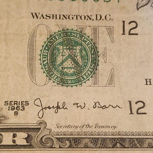May include: Close-up of a one-dollar bill. The bill features the text "WASHINGTON, D.C." and the number "12". The bill also includes the text "SERIES 1963 B" and the signature of Joseph W. Barr, Secretary of the Treasury.