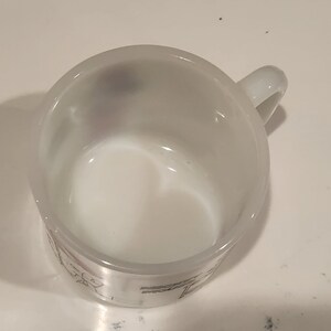 May include: White glass mug with a handle. The mug has a cylindrical shape and a clear handle. The inside of the mug is white. The bottom of the mug has a design with text.