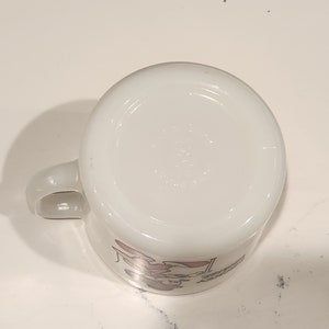 May include: White ceramic mug with a handle, featuring a cartoon character design in pink and red. The mug is viewed from the bottom, showing the base and a portion of the design. The mug is on a white surface.