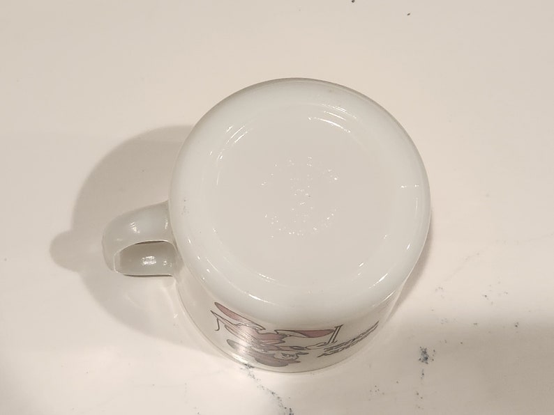 May include: White ceramic mug with a handle, featuring a cartoon character design in pink and red. The mug is viewed from the bottom, showing the base and a portion of the design. The mug is on a white surface.