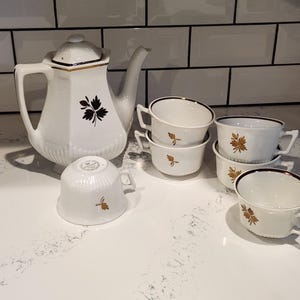 May include: A white porcelain tea set with a teapot and several teacups. The teapot has a black floral design and gold trim. The teacups have a brown leaf design. The set is on a white countertop.