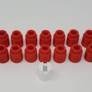 Könnte beinhalten: Ein Satz von 12 roten Plastikkappen mit den Nummern 17 bis 28 darauf gedruckt. Die Kappen sind in zwei Reihen zu sechs angeordnet. Eine weiße Plastikkappe mit einem Schlitz befindet sich in der Mitte des Bildes.