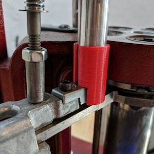 May include: A close-up of a reloading press with a red 3D printed sleeve on the ram. The press is made of metal and has a silver finish.