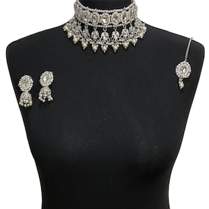 以下が含まれることがあります： チョーカーネックレス、イヤリング、ヘッドピースを備えた、シルバーカラーのブライダルジュエリーセット。 ネックレスとヘッドピースには、透明な楕円形の石と小さな揺れるパールが付いています。 イヤリングは、ベル型のデザインです。