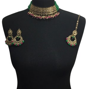 May include: A gold necklace set with green and pink beads, a matching pair of earrings, and a matching maang tikka. The necklace is a choker style with a large pendant in the center.