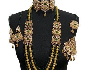 Op de afbeelding: Een gouden en groene bruidssieradenset met een chokerketting, een lange gelaagde ketting, oorbellen en een maang tikka. De set heeft ingewikkelde ontwerpen met groene stenen en parels.