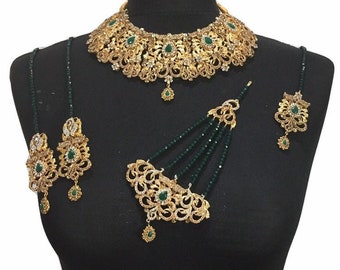 Op de afbeelding: Een gouden en groene bruidssieradenset met een ketting, oorbellen, een maang tikka en een armband. De set is versierd met ingewikkelde ontwerpen en groene stenen.
