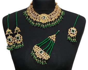 Op de afbeelding: Een Indiase bruidssieradenset van goud en groen met een ketting, oorbellen, een maang tikka en een neusring. De set heeft ingewikkelde bloemmotieven en groene stenen.