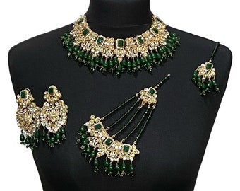 Op de afbeelding: Een goudkleurige sieradenset met een ketting, oorbellen, een hoofdband en een haaraccessoire. De set bevat smaragdgroene edelstenen en bungelende kralen. De sieraden worden getoond op een zwarte mannequin.