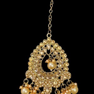 Pode incluir: Pe&ccedil;a de cabe&ccedil;a dourada com um pendente em forma de gota. O pendente &eacute; adornado com pequenas pedras preciosas transparentes e uma p&eacute;rola central. P&eacute;rolas adicionais pendem da parte inferior, suspensas por delicadas correntes. Uma corrente estende-se para cima para prender a pe&ccedil;a.