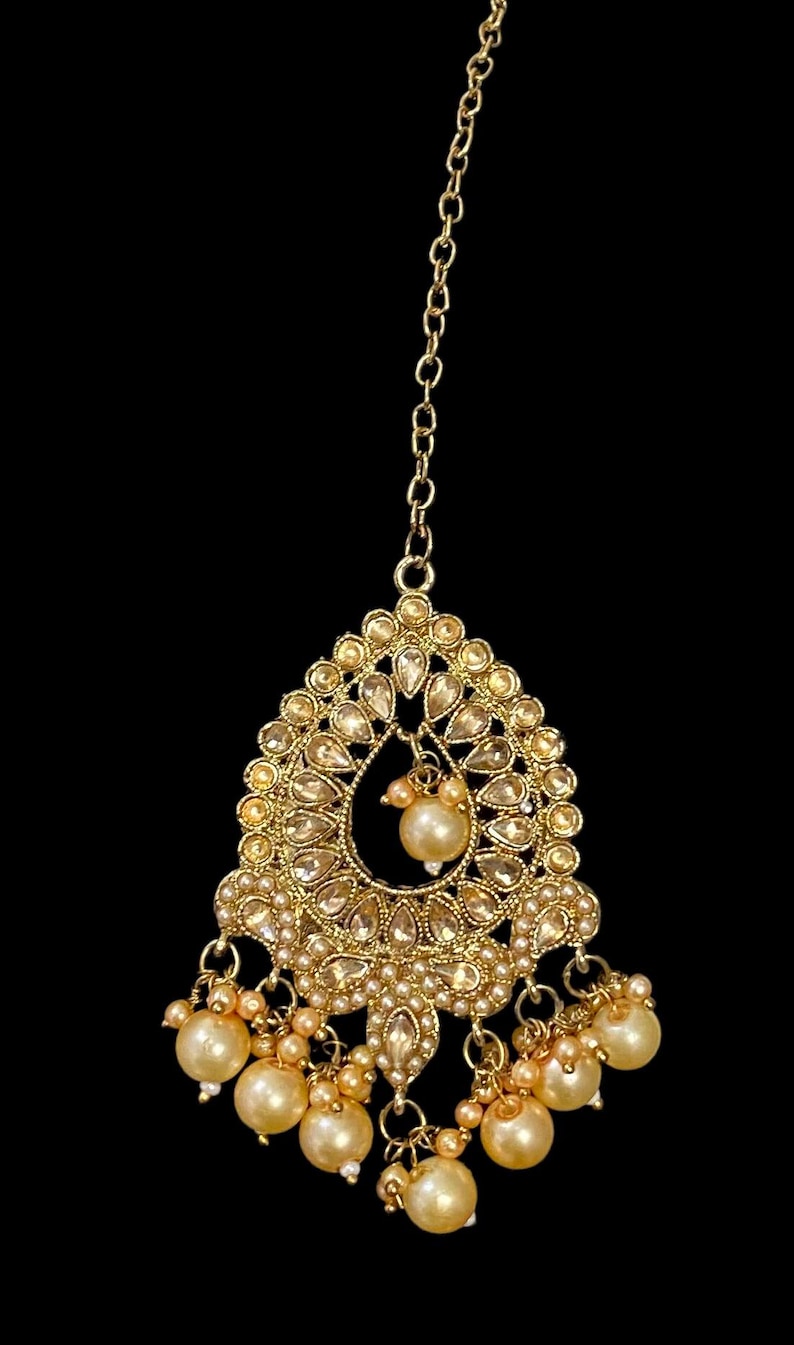 Pode incluir: Pe&ccedil;a de cabe&ccedil;a dourada com um pendente em forma de gota. O pendente &eacute; adornado com pequenas pedras preciosas transparentes e uma p&eacute;rola central. P&eacute;rolas adicionais pendem da parte inferior, suspensas por delicadas correntes. Uma corrente estende-se para cima para prender a pe&ccedil;a.