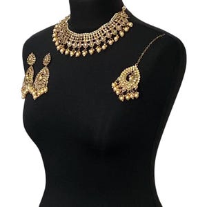 Pode incluir: Um conjunto de joias douradas exibido em um manequim preto. O conjunto inclui um colar, brincos e uma pe&ccedil;a de cabe&ccedil;a. O colar e a pe&ccedil;a de cabe&ccedil;a apresentam designs intrincados com pequenos detalhes redondos semelhantes a p&eacute;rolas. Os brincos t&ecirc;m um design semelhante.