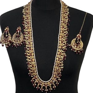 Könnte beinhalten: Ein goldfarbenes Halsketten-Set mit passenden Ohrringen und einem Kopfschmuck. Der Schmuck hat bordeauxrote und weiße Akzente. Die lange Halskette wird auf einer schwarzen Schaufensterpuppe präsentiert.
