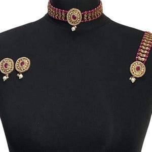 May include: A three-piece jewellery set displayed on a black mannequin. The set includes a choker necklace, a bracelet, and a pair of earrings. Each piece features a maroon centre stone surrounded by gold-coloured metal and small white pearls.