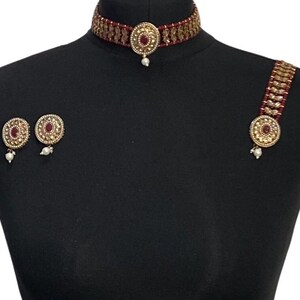 Op de afbeelding: Een goudkleurige sieradenset met een choker-ketting, oorbellen en een armband. De set bevat een choker-ketting, oorbellen en een armband. Elk stuk heeft een centrale rode steen en is geaccentueerd met kleinere goudkleurige details en parels.