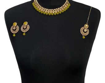 May include: A gold-toned jewelry set displayed on a black mannequin. The set includes a necklace, earrings, and a headpiece. The necklace and earrings feature yellow-green beads and small white pearls. The headpiece has a similar design, with a dangling yellow-green bead.
