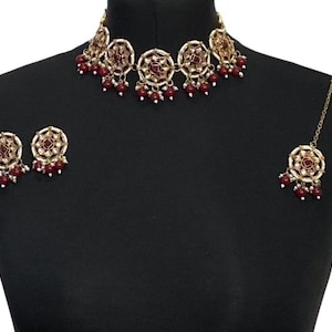 Könnte beinhalten: Ein goldfarbenes Schmuckset mit Halskette, Ohrringen und Kopfschmuck. Die Teile haben ein kreisförmiges Design mit roten und weißen Akzenten und kleinen roten Perlenanhängern. Der Schmuck wird auf einer schwarzen Schaufensterpuppe präsentiert.