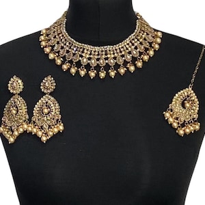 Pode incluir: Um conjunto de joias douradas exibido em um manequim preto. O conjunto inclui um colar gargantilha, um par de brincos e um adorno de cabe&ccedil;a. O colar e os brincos apresentam um design floral com pequenos detalhes de p&eacute;rolas. O adorno de cabe&ccedil;a tem um design semelhante e gotas de p&eacute;rolas.