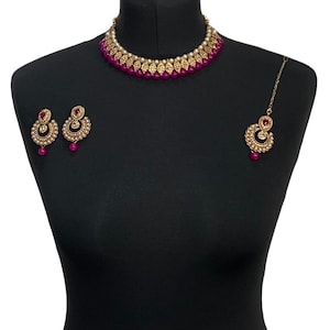 May include: A gold-coloured jewellery set comprising a necklace, earrings, and a headpiece. The necklace and earrings feature a design with small, round, magenta-coloured beads. The headpiece has a similar design. The jewellery is displayed on a black mannequin.
