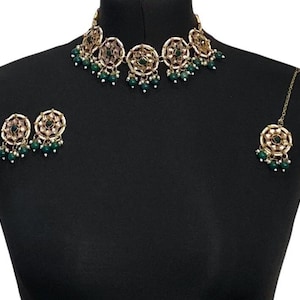 May include: A gold-toned jewelry set featuring a choker necklace, earrings, and a headpiece. The necklace and earrings have circular designs with green and pink accents, and dangling green beads. The headpiece has a similar design.