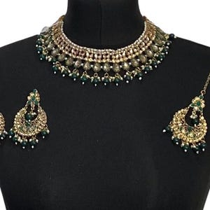 Op de afbeelding: Een goudkleurige sieradenset met een ketting, oorbellen en een hoofdband. De ketting en oorbellen hebben een bloemmotief met groene en witte accenten. De hoofdband heeft een druppelvorm.