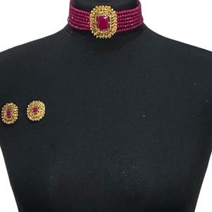 Op de afbeelding: Een bordeauxrode kralen choker ketting met een goudkleurig middelpunt en bijpassende oorstekers. Het middelpunt van de ketting en oorbellen heeft een rechthoekige bordeauxrode steen. De sieraden worden getoond op een zwarte mannequin.