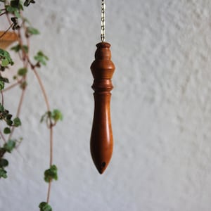 Puede incluir: Un péndulo de madera con una cadena dorada. El péndulo es marrón y tiene una punta puntiaguda.