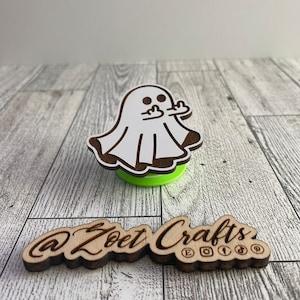 May include: A wooden ghost-shaped decoration with a white body and brown outline, giving the middle finger. The ghost is on a green base. Below it is a wooden sign with the text "@Zoel Crafts" and social media icons.