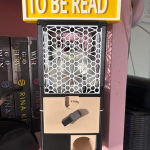 May include: A black, white, and beige plastic dispenser featuring a yellow sign at the top with the text TO BE READ LN. The unit has a white geometric lattice front, a beige front panel with a black turning knob, and a small circular opening at the base.