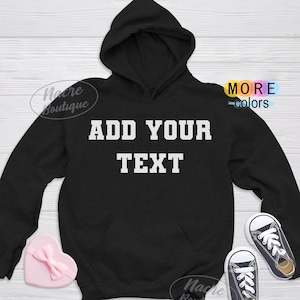 May include: Black hooded sweatshirt with the text "ADD YOUR TEXT" in white block letters. The sweatshirt has a front pocket and a hood. A pink heart-shaped box and a pair of small black sneakers are also in the image.