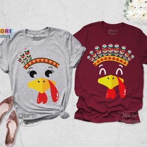 May include: Two short-sleeved t-shirts, one gray and one burgundy, feature a cartoon turkey face design. The turkey has a yellow beak, red wattle, and a colorful headdress. The text "MORE colors" is visible on the left.