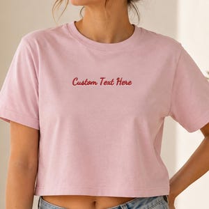 May include: A light pink short-sleeved crop top featuring the embroidered red text Custom Text Here centered on the chest. The garment has a crew neckline and a straight hemline, constructed from a solid-colored fabric with a smooth texture.