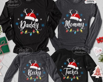 Puede incluir: Camisetas negras y grises de manga larga con diseños navideños. Cada camiseta presenta un gorro de Papá Noel con cuernos, los nombres "Daddy", "Mommy", "Becky" o "Tucker", copos de nieve y luces de cadena. Las camisetas son de Nacre Boutique.