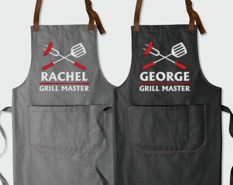 May include: Two grey and black denim aprons with adjustable straps. The aprons have a large pocket on the front and an embroidered design of crossed grilling tools with a hot dog and the text "Grill Master". One apron says "Rachel" and the other says "George".