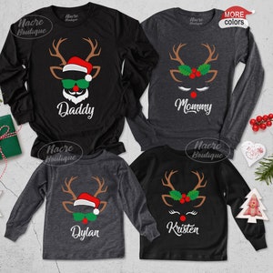Pode incluir: Quatro camisas de Natal de manga comprida com desenhos de renas. As camisas s&atilde;o pretas e cinzentas, com as palavras "Daddy", "Mommy", "Dylan" e "Kristen" impressas sob as caras das renas. As renas t&ecirc;m chifres, &oacute;culos de sol e gorros de Pai Natal.