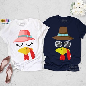 May include: Two short-sleeved t-shirts with turkey face designs. One is white with a pink hat and blue bow, the other is navy blue with a brown hat and sunglasses. Both shirts have a yellow beak and red wattle.