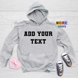 May include: A light gray hoodie with a front pocket and hood. The text "ADD YOUR TEXT" is printed in black block letters across the chest. A pink heart-shaped box and a pair of black and white sneakers are next to the hoodie.