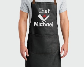 May include: Black denim apron with adjustable brown straps. The apron has a large pocket and says "Chef Michael" with a crossed knife and flame graphic.