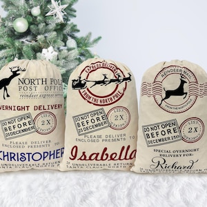 May include: Three white canvas Santa sacks with red and black printed designs. The sacks have the text "North Pole Post Office", "Express Delivery from the North Pole", and "Reindeer Mail" printed on them. Each sack has a personalized name: Christopher, Isabella, and Richard.