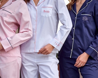 May include: Three women wearing matching pajamas. The bride's pajamas are white with pink piping and the text "Bride" embroidered on the chest. The other two pajamas are pink and navy blue with white piping and monogrammed with the initials "CBS".