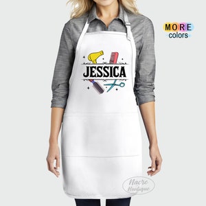 Puede incluir: Delantal blanco con borde negro y dos bolsillos. El delantal tiene un dise&ntilde;o gr&aacute;fico de un secador de pelo, un peine, unas tijeras y la palabra "JESSICA" en texto negro.