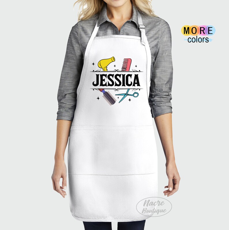 Puede incluir: Delantal blanco con borde negro y dos bolsillos. El delantal tiene un dise&ntilde;o gr&aacute;fico de un secador de pelo, un peine, unas tijeras y la palabra "JESSICA" en texto negro.