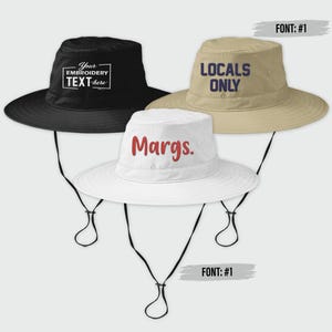 May include: Three bucket hats with chin straps. One black hat has the text "Your EMBROIDERY TEXT here". A tan hat has the text "LOCALS ONLY". A white hat has the text "Margs." All hats have a wide brim.
