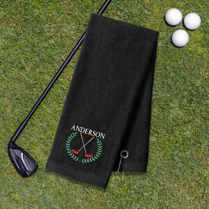 A black towel with a personalized name and logo, a golf club, and three golf balls on a grassy surface.