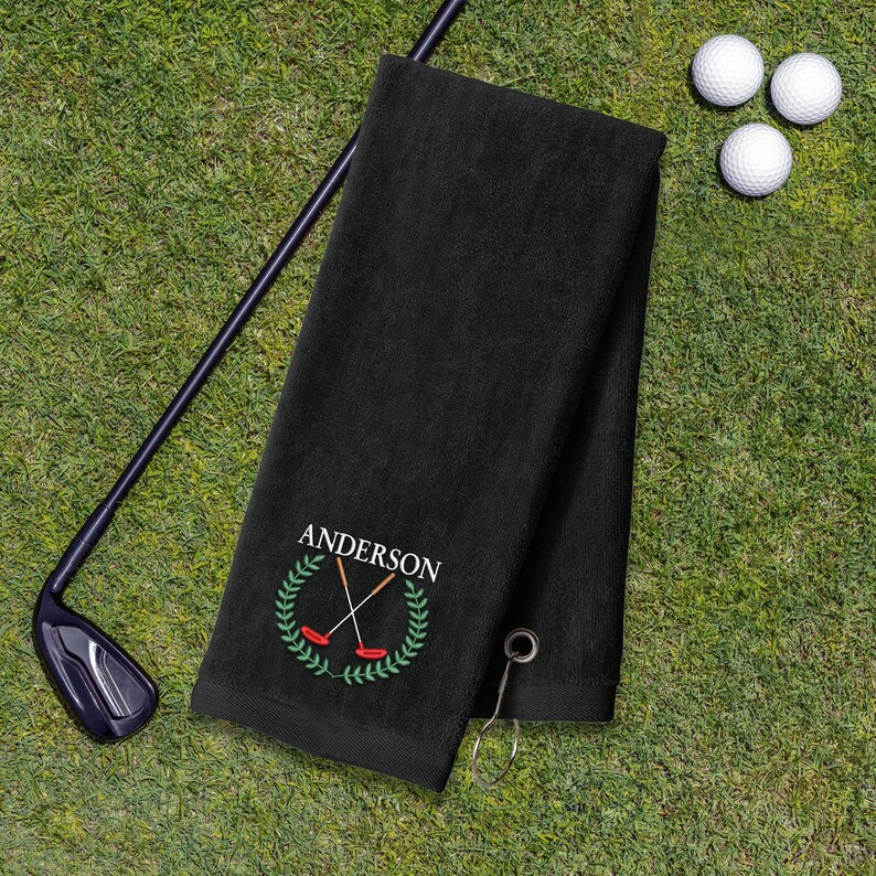 A black towel with a personalized name and logo, a golf club, and three golf balls on a grassy surface.