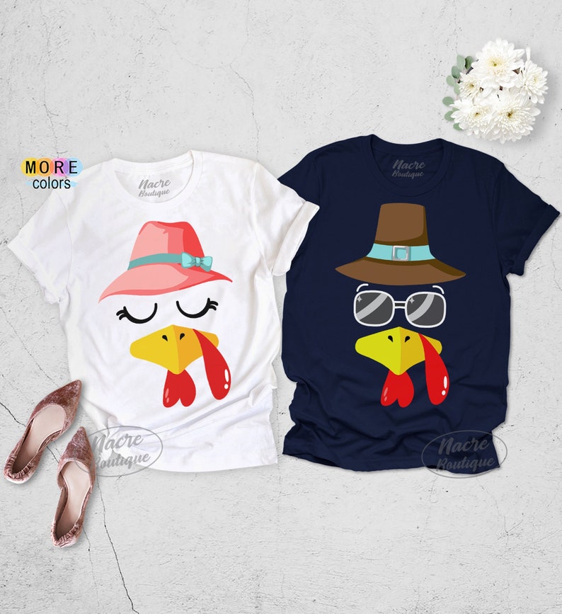 May include: Two short-sleeved t-shirts with turkey face designs. One is white with a pink hat and blue bow, the other is navy blue with a brown hat and sunglasses. Both shirts have a yellow beak and red wattle.