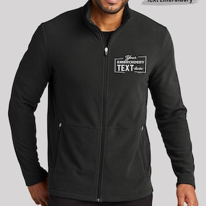 May include: Black zip-up fleece jacket with a high collar and two side pockets. The jacket has a white embroidered text box that says "Your Embroidery TEXT here" and "Text Embroidery" above the model's head.
