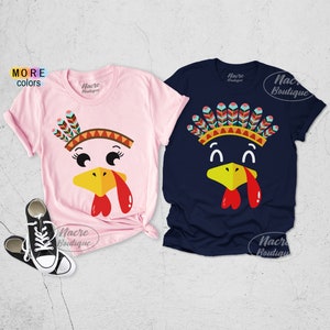 May include: Two short-sleeved t-shirts with turkey face designs. One is light pink, the other is navy blue. Each shirt features a turkey face with a yellow beak, red wattle, and a Native American headdress. The brand name "Nacre Boutique" is printed on the shirts.
